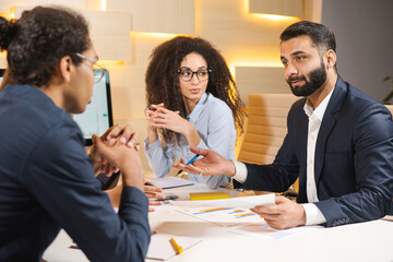 Listen attentively. Skillful young pleasant businessman in shirt is sitting at table and holding pen while looking at responsible colleague male who sitting in front of him