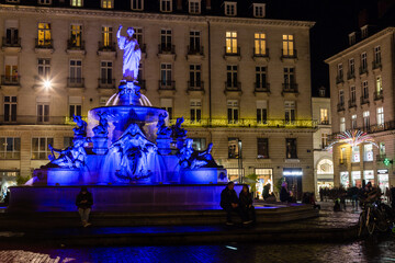 Place d'une ville avec fontaine du 19e siècle, de nuit éclairée en bleu avec personnes autour....