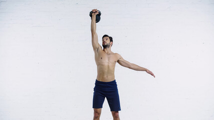 muscular sportsman holding heavy kettlebell above head near white brick wall - Powered by Adobe