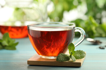 Glass cup of aromatic black tea with fresh mint on light blue wooden table against blurred background, closeup