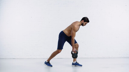 full length of muscular sportsman in sneakers exercising with heavy kettlebell near white brick wall