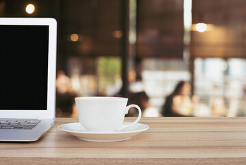 Laptop and white coffee mug placed on wooden table in coffee shop with people sitting and drinking coffee in the restaurant at morning before start work.