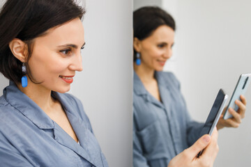 Obraz premium Bruinette woman in pajama working online with smartphone in hands at home. Female using phone looking at screen typing message, using telephone technology doing online job, surfing internet social