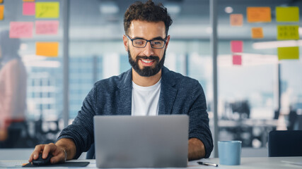 Modern Office: Portrait of Young Stylish Businessman Wearing Glasses Works on Laptop, Does Data...