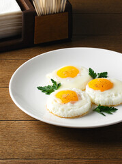 Fried eggs for breakfast. Fried eggs on a white plate on a wooden table. Parsley, dill. Breakfast at the hotel, cafe, restaurant. Copy space. Morning meal. Space for text. Food photography. Three eggs