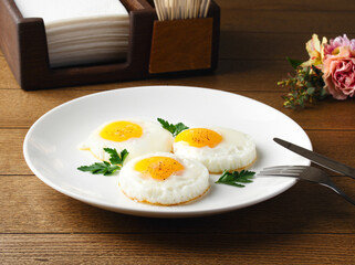 Fried eggs for breakfast. Fried eggs on a white plate on a wooden table. Parsley, dill. Breakfast at the hotel, cafe, restaurant. Copy space. Morning meal. Space for text. Food photography. Three eggs