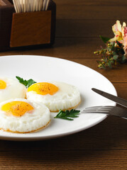 Fried eggs for breakfast. Fried eggs on a white plate on a wooden table. Parsley, dill. Breakfast at the hotel, cafe, restaurant. Copy space. Morning meal. Space for text. Food photography. Three eggs