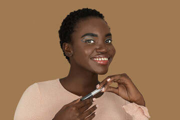 Closeup portrait of a smiling young black woman with short Afro hair, light makeup and lipstick posing by herself holding a lipstick in both of her hands inside a studio with a pecan background.