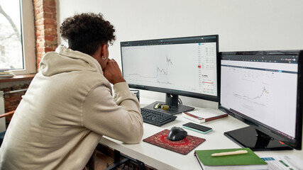 Rear view of young guy trader looking at computer screen to analyze statistics while sitting at home office desk