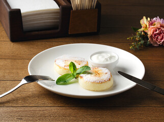 Russian cheesecakes on a white plate with cream on wooden background. Breakfast at the hotel, cafe, restaurant. Copy space. Morning meal. Space for text. Food photography. Two cheesecakes with mint