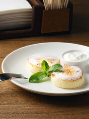 Russian cheesecakes on a white plate with cream on wooden background. Breakfast at the hotel, cafe, restaurant. Copy space. Morning meal. Space for text. Food photography. Two cheesecakes with mint