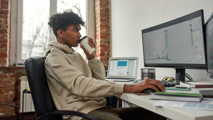 Side view of young male trader drinking coffee, working on computer, studying charts while trading from home