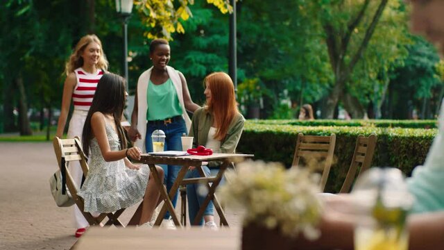 In the middle of the park at the cafe beautiful two ladies have a conversation while other ladies black woman and blonde hair lady came to their friends they hugging each other and enjoy the time