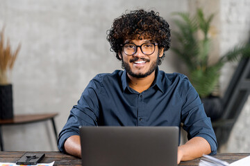 Portrait of smiling and friendly multi ethnic male freelancer in glasses using laptop and looks at the camera, indian latin curly guy in smart casual shirt typing, working remotely, develops software