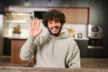 Portrait of happy young Indian guy sitting at desk waving at camera, smiling curly ethnic man in casual hoodie involved virtual meeting, hispanic male student taking a part in educational conference