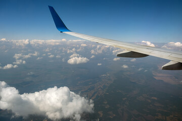 View of jet airplane wing from inside flying over white puffy clouds in blue sky. Travel and air transportation concept