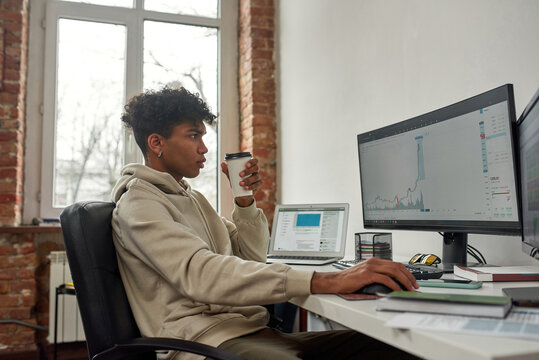 Side view of young male trader drinking coffee, working on computer, studying charts while trading from home
