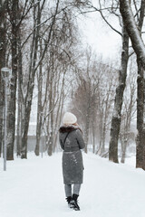 Fototapeta na wymiar Back view of young woman in coat and knitted white hat enjoying walk in winter park, outdoors. Rear view of lonely girl walking along snowy alley. Vertical image