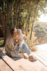 Happy smiling mother wear trench coat playing with baby boy 1 year old wear knit suit clothes sitting on wooden pier over lake nature background and sun light. Spring or autumn season. Motherhood.