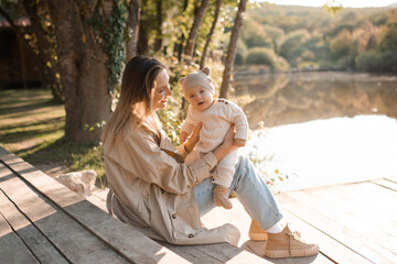 Happy smiling mother wear trench coat playing with baby boy 1 year old wear knit suit clothes sitting on wooden pier over lake nature background and sun light. Spring or autumn season. Motherhood.