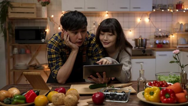 asian romantic couple looking at recipes on tablet computer while having discussion about what to cook for valentine&rsquo;s day dinner at home. the man propping his head while thinking