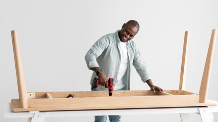 Happy african american handyman drilling wooden planks of table, assembling new furniture with...