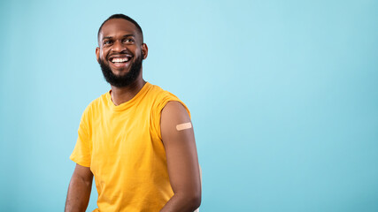 Cheerful vaccinated black man with plaster bandage on his shoulder after covid-19 vaccine injection on blue background - obrazy, fototapety, plakaty