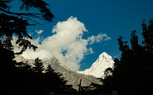 majestic himalayan mountain Mt sudarshan covered in snow. silhouetted tree line in foreground. blue sky and drifting clouds.