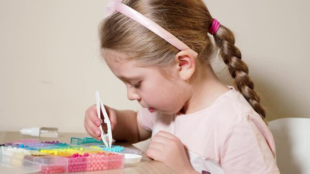 A little preschool girl plays aquamosaic. The child makes up blue plastic balls with tweezers on a stencil. Side view. Multi-colored balls. The concept of children's leisure and self-development