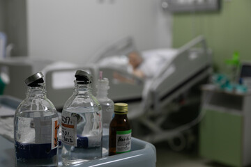 Glass bottles on a medical solution stand on the medical table in focus in the foreground. In the background, a seriously ill patient is lying on a bed in a hospital room of an intensive care unit.