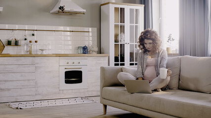 Pregnant freelancer holding cup and using laptop on couch at home