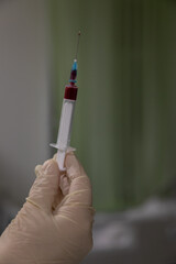 Medic's hand in a white latex glove holds a small 5ml syringe filled with red blood. There is a...