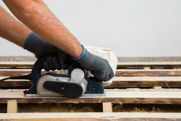 Young adult man hands in protective gloves using jointer and shaving old wooden floor planks. Closeup. Side view. Preparing for repair work of home. Flooring restoration.
