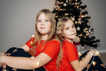 Teenage sisters in red holiday T-shirts sitting at the Christmas tree and celebrating the New Year