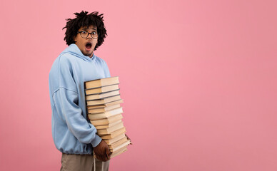 Nerdy black teenage student feeling shocked over pile of homework, holding big stack of books on...
