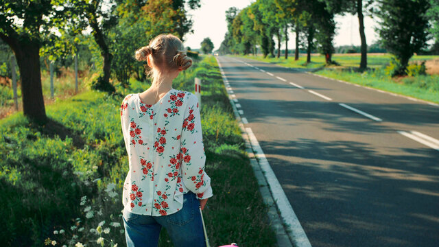 Young woman on countryside road. Back view of young female with red suitcase standing on roadside while travelling through countryside.