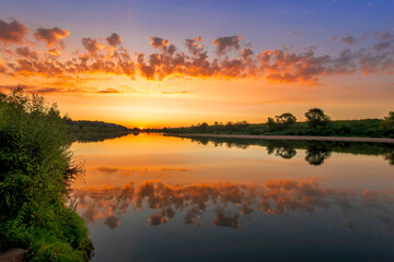 Naklejka na ściany i meble Amazing view at scenic landscape on a beautiful river and colorful sunset with reflection on water surface and glow on a background, spring season landscape