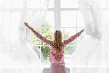 Young pretty blonde woman in pink pajamas woke up, look at large panoramic window in bedroom