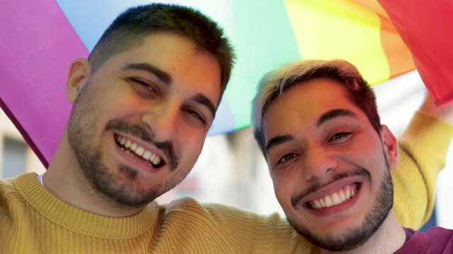 Young gay male couple under LGBT rainbow flag at pride parade event