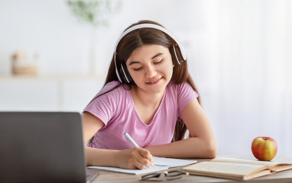 Home Schooling. Joyful Indian Teenage Girl In Headphones Taking Notes During Online Class On Laptop Indoors