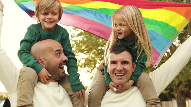 Gay male couple with children having fun holding LGBT rainbow flag outdoor at city park - Family and love concept