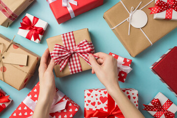 First person top view photo of valentine's day decor presents and young woman's hands unwrapping craft paper giftbox with checkered ribbon bow on isolated pastel blue background
