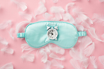 Top view photo of white alarm clock on blue silk sleeping mask and feathers on isolated pastel pink background