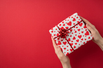 First person top view photo of saint valentine's day decorations female hands holding giftbox in white wrapping paper with heart pattern and red star bow on isolated red background with empty space