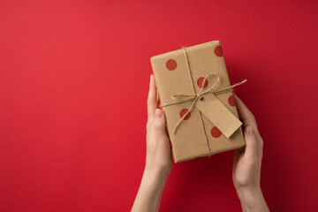 First person top view photo of st valentine's day decorations female hands demonstrating kraft paper giftbox with polka dot pattern and twine bow on isolated red background with copyspace