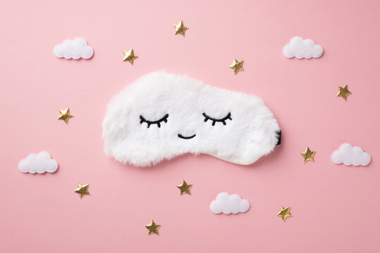 Top view photo of white fluffy sleeping mask with closed eyes and smile clouds and golden stars on isolated pastel pink background