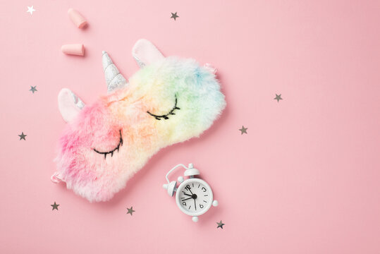 Top view photo of funny multicolored fluffy unicorn sleeping mask small white alarm clock earplugs and star shaped confetti on isolated pastel pink background with copyspace