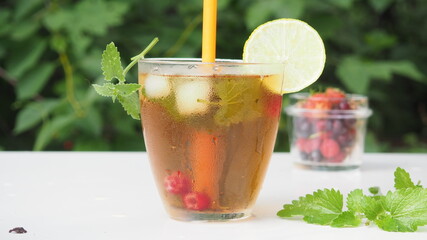 Fototapeta premium Iced fruit tea or cold berry drink in clear glass with mint leaf. fresh berries raspberries, blueberries, strawberries, lime. Refreshing summer drink. nature foliage background