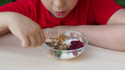 boy eats proper nutrition and healthy breakfast bowl of oat granola with yogurt, fresh raspberries, blueberries, strawberries, blackberries and nuts with spoon on white for healthy breakfast, top view