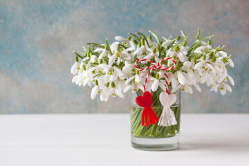 A bouquet of snowdrops in a glass on a table, a red and white symbol of a martenitsa and a heart on...